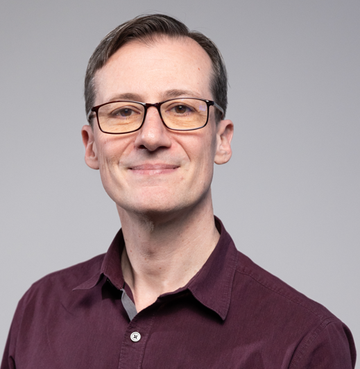 A man with short dark hair, wearing glasses, and a maroon button-up shirt, smiling at the camera against a plain gray background.