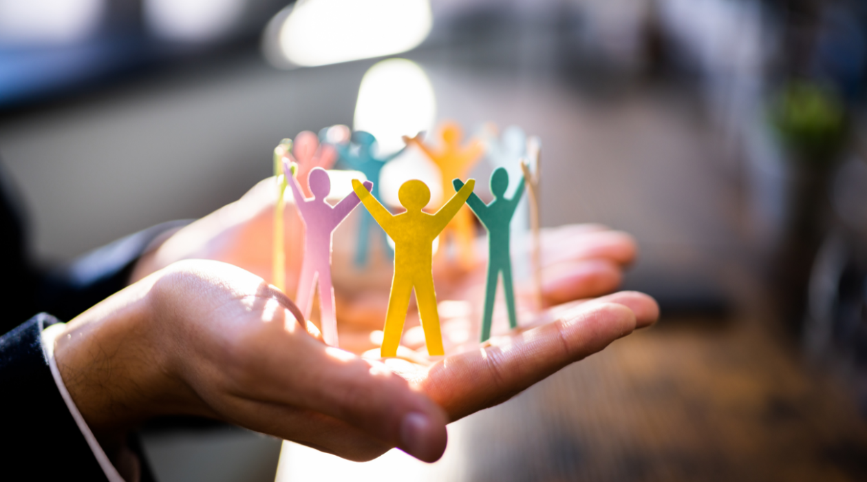 Hand holding paper cutouts of multicolored human figures with arms raised, arranged in a circle.
