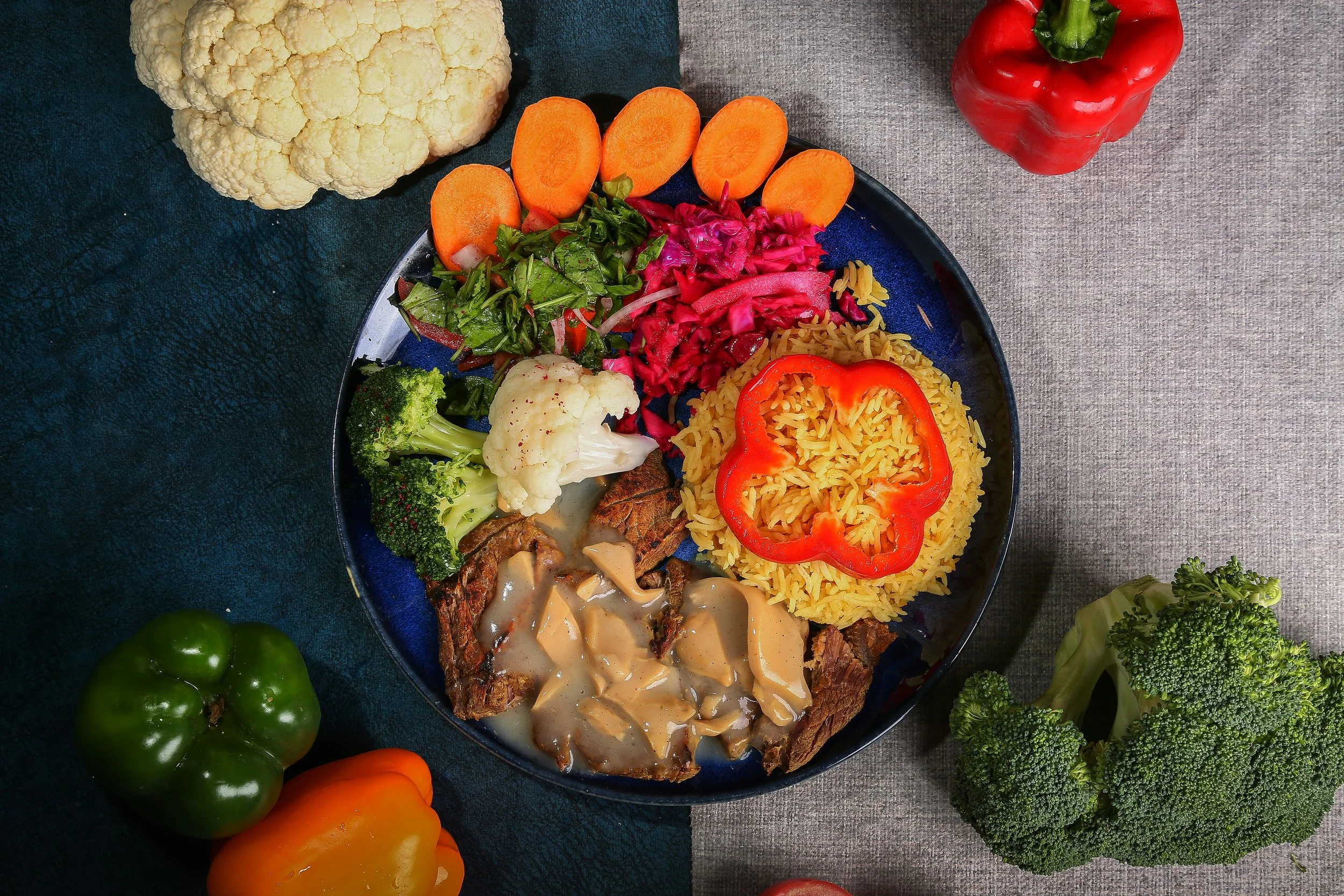 Plate of colorful Mexican food with rice, beef, vegetables, and sauces, surrounded by fresh vegetables including cauliflower, broccoli, bell peppers, carrots, and tomatoes.
