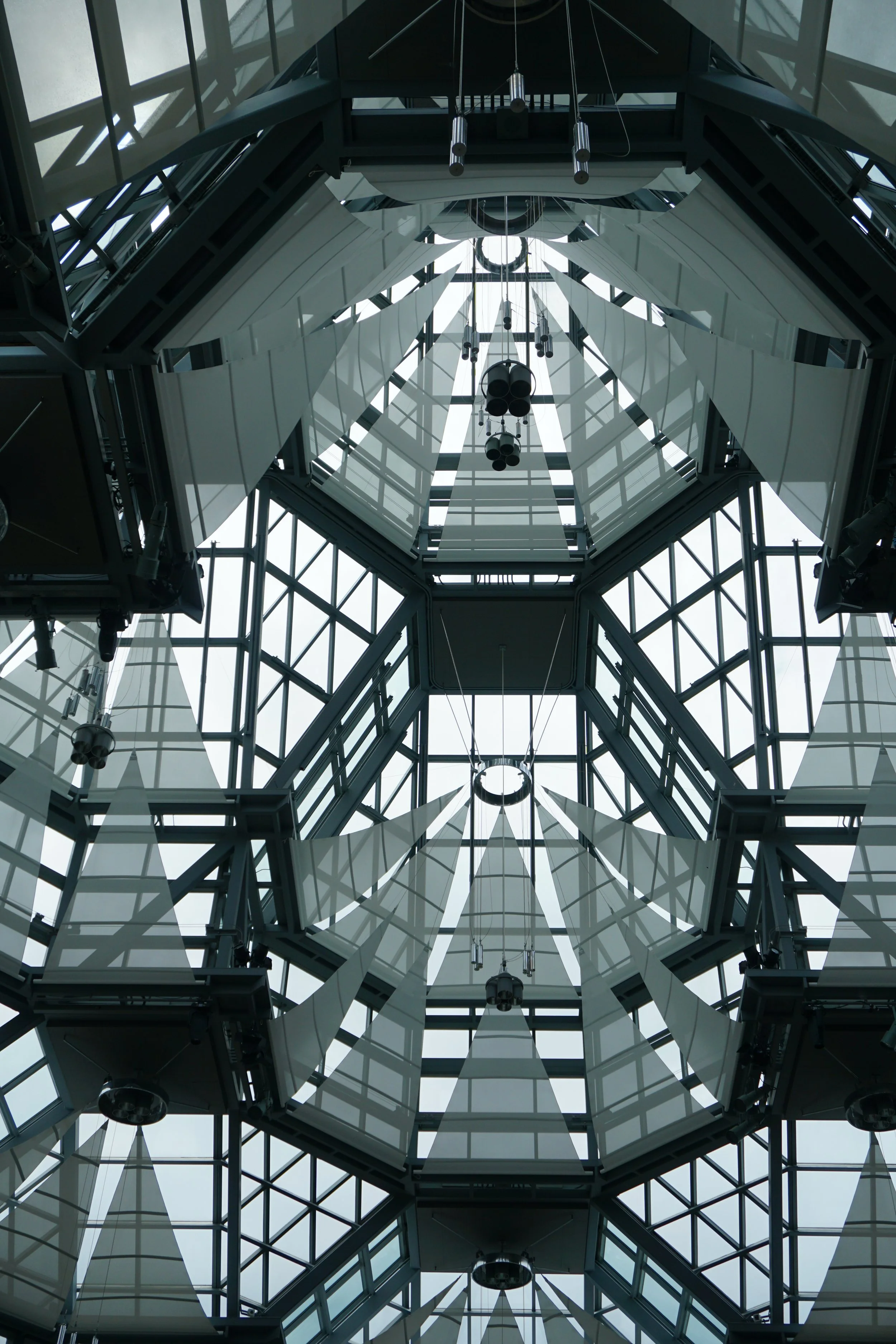 Looking up at the geometric glass and metal ceiling of a modern building with hanging lights.