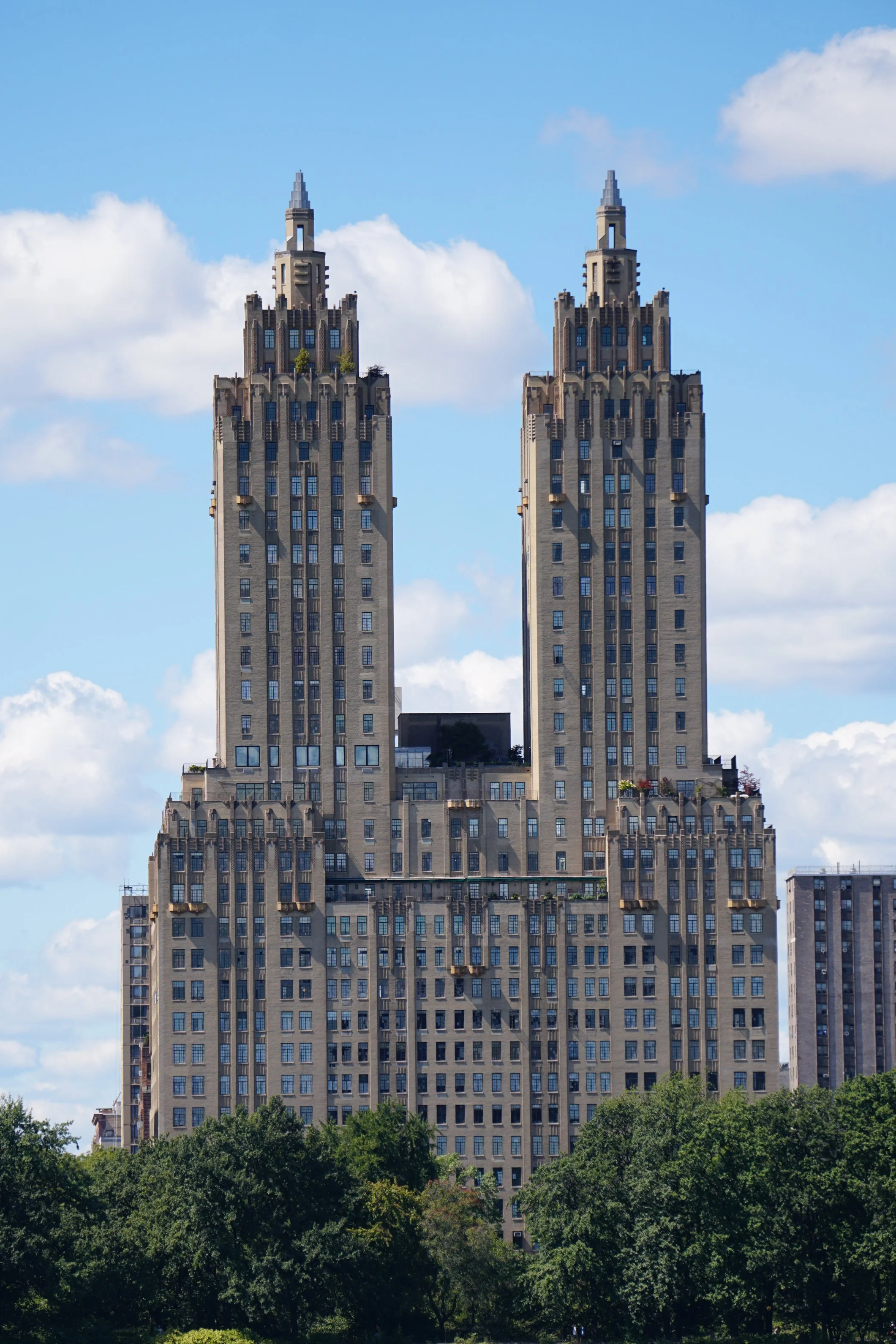 Two tall, identical skyscrapers with spires on top, set against a blue sky with scattered clouds. The buildings are beige with many windows and some greenery and rooftops at the lower levels, surrounded by trees.