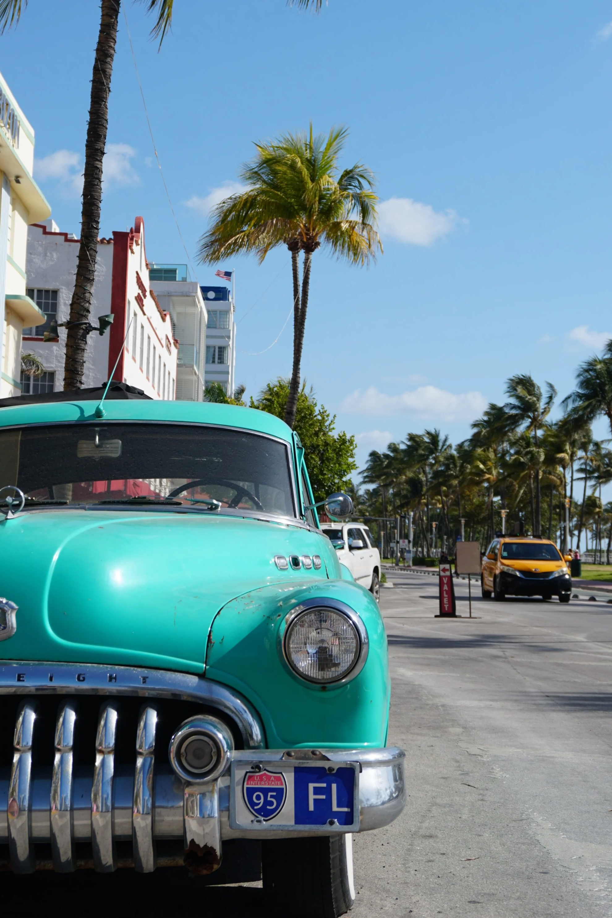 A vintage teal car parked along a street in a coastal area with palm trees, modern buildings, and a bright blue sky in the background.