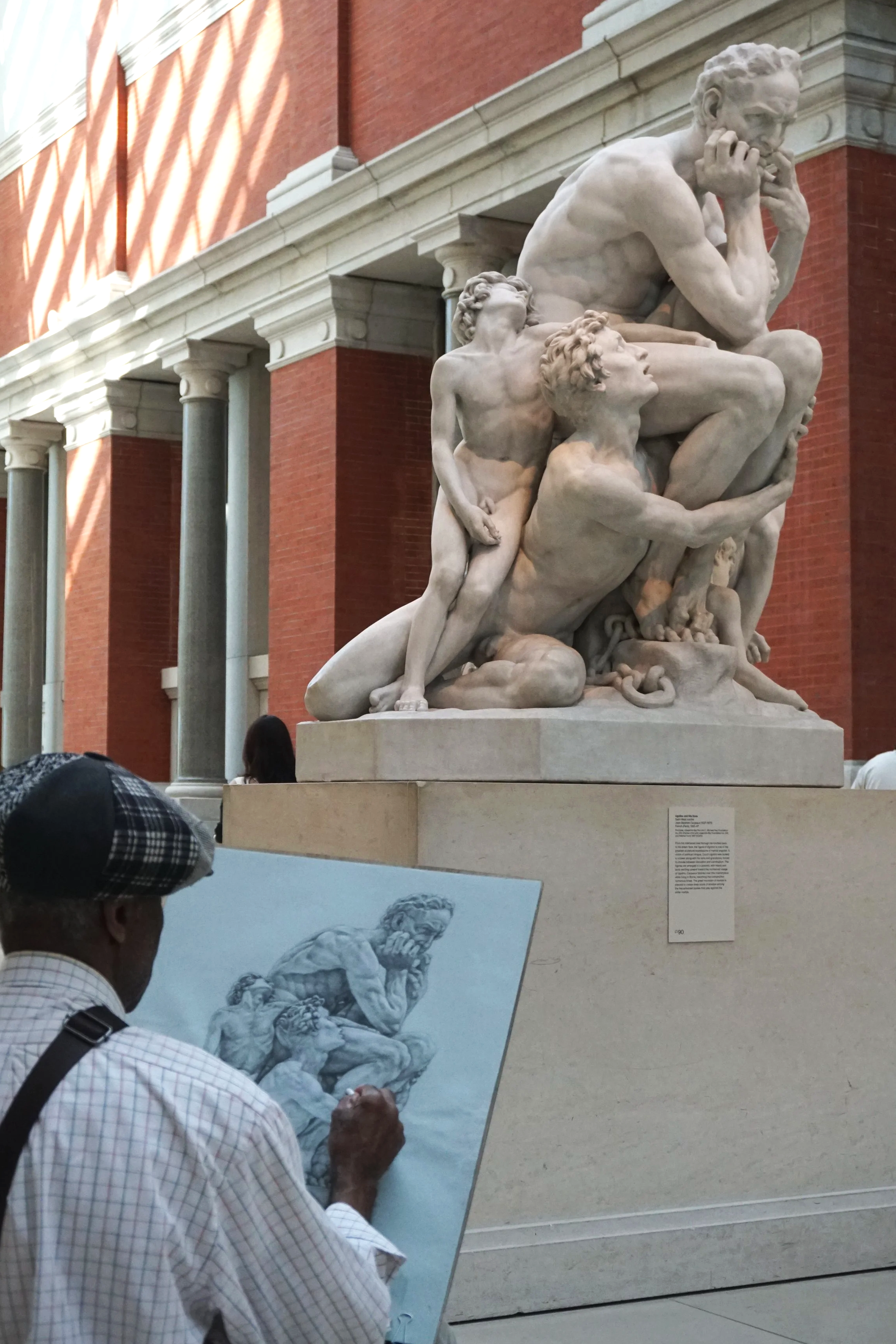 A man sketching a marble sculpture of multiple figures displayed in a museum.