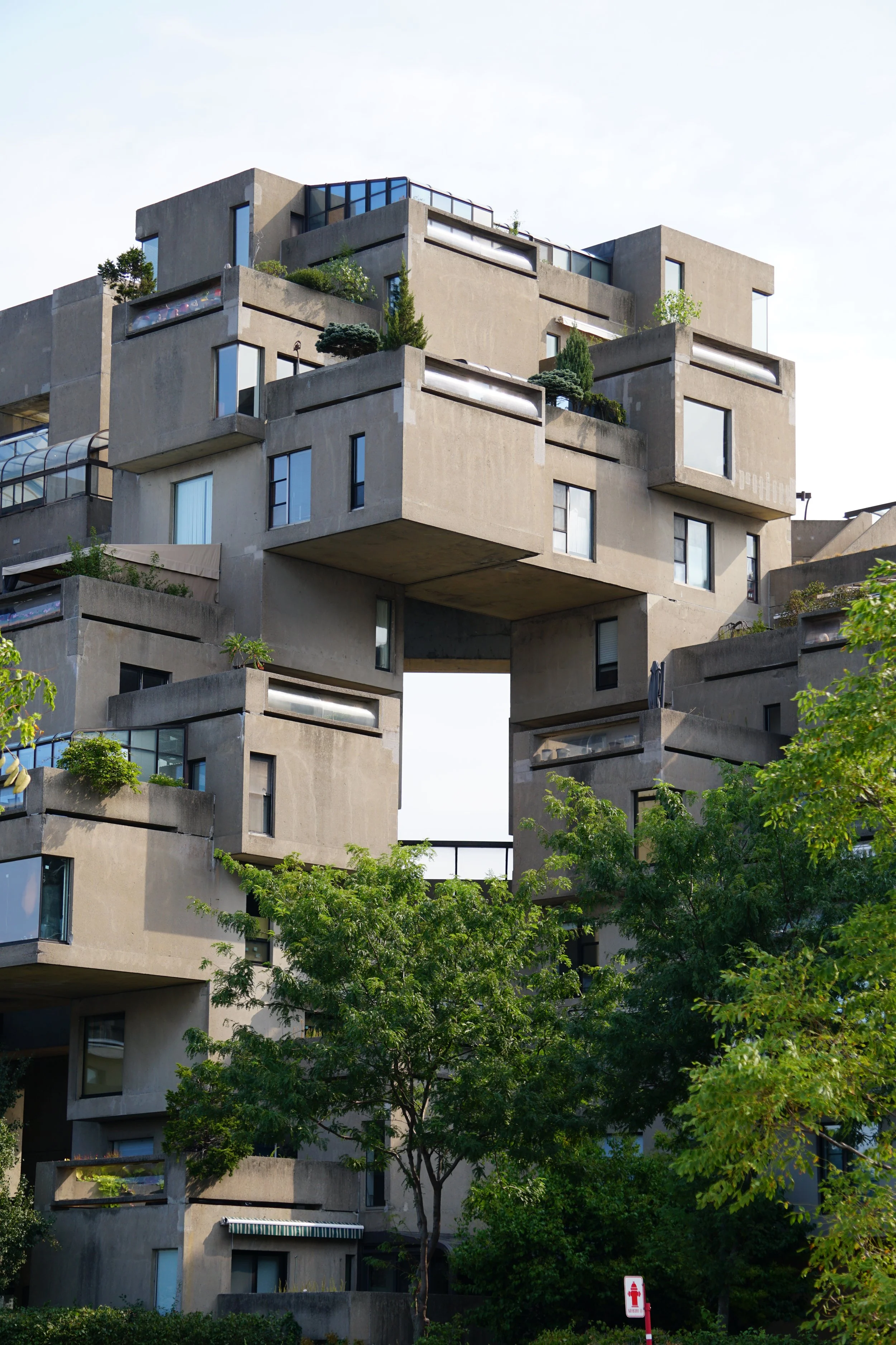 Modern multi-story apartment building with an unconventional design, featuring stacked concrete blocks with windows and small plants, surrounded by trees.