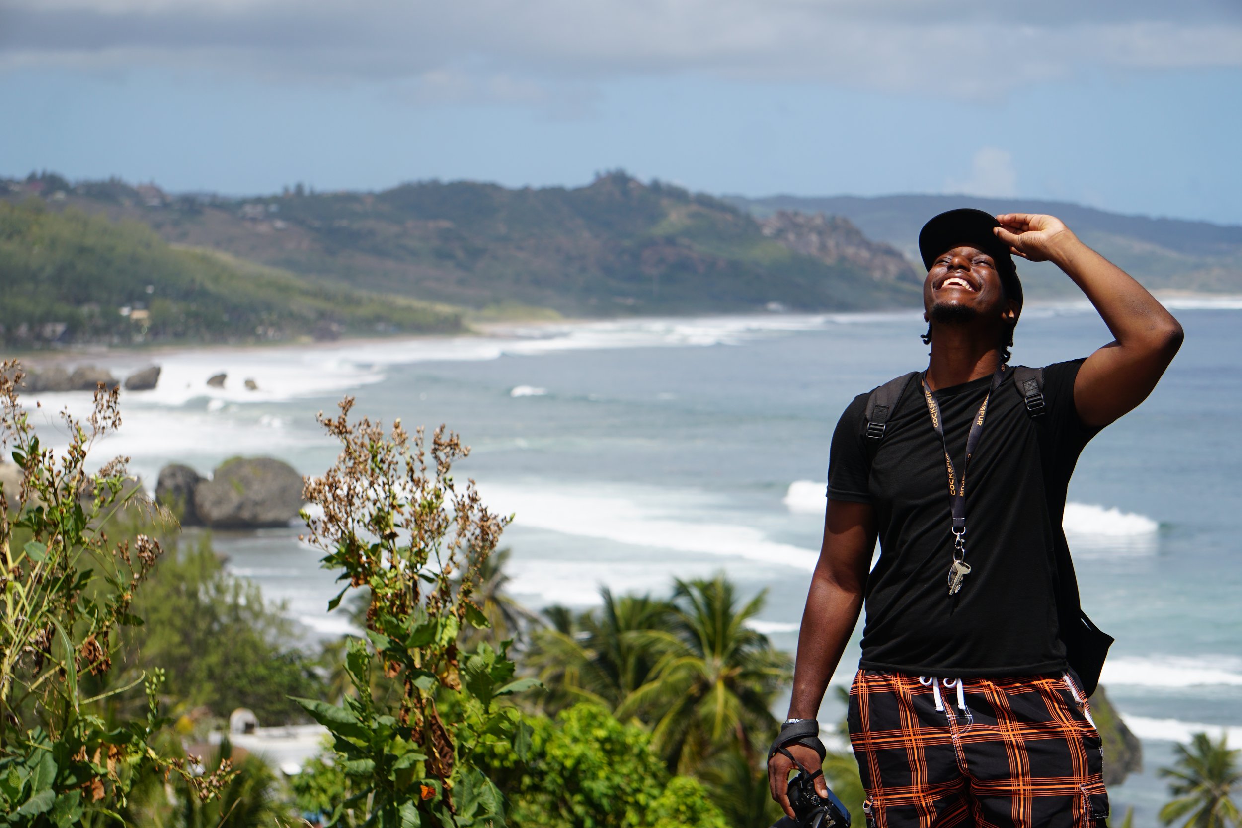 Man in black shirt and plaid shorts smiling and shielding eyes while standing outdoors with a beach, ocean, and hills in the background.
