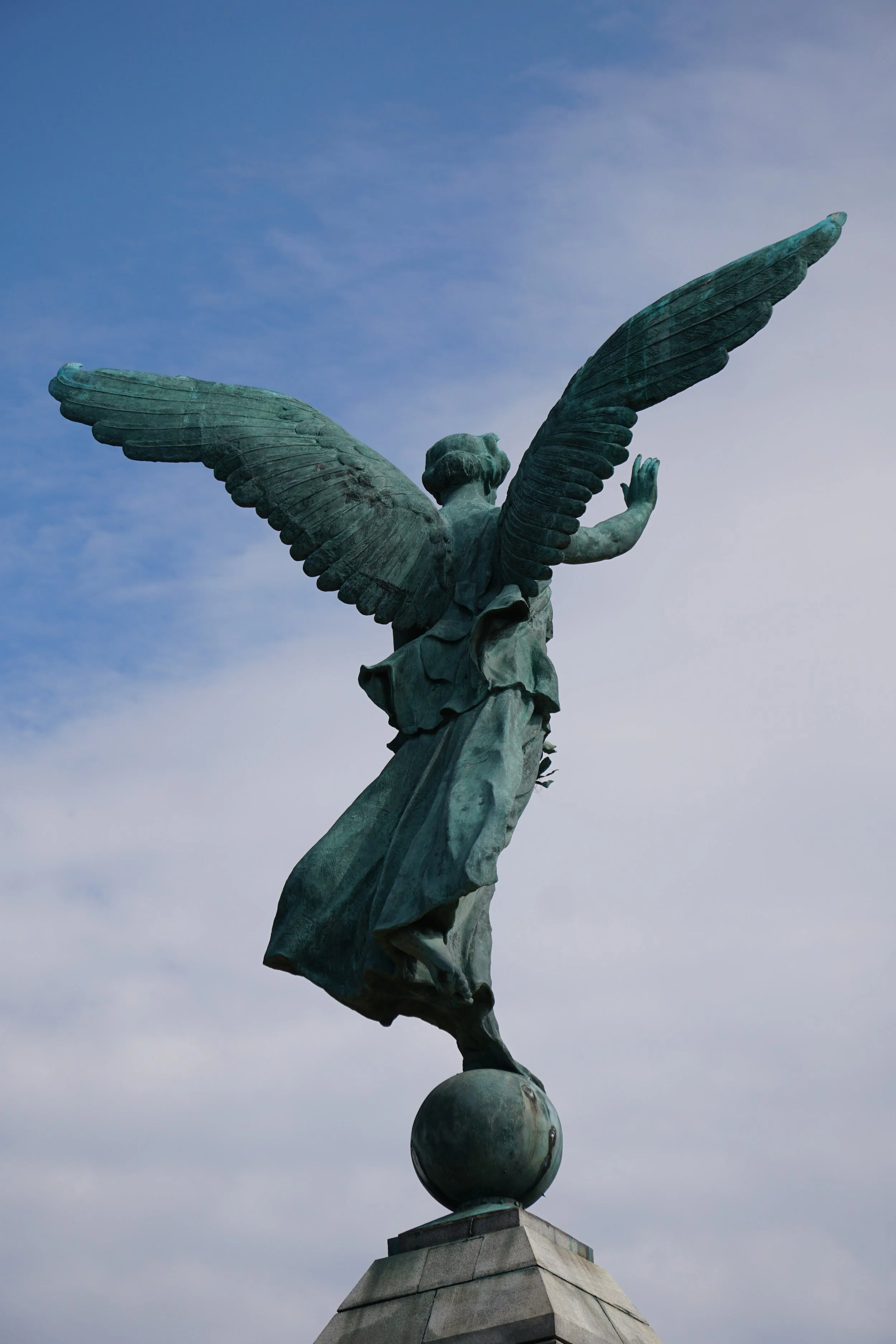 Bronze statue of a winged woman standing on a globe, extending one hand forward, set against a cloudy sky.