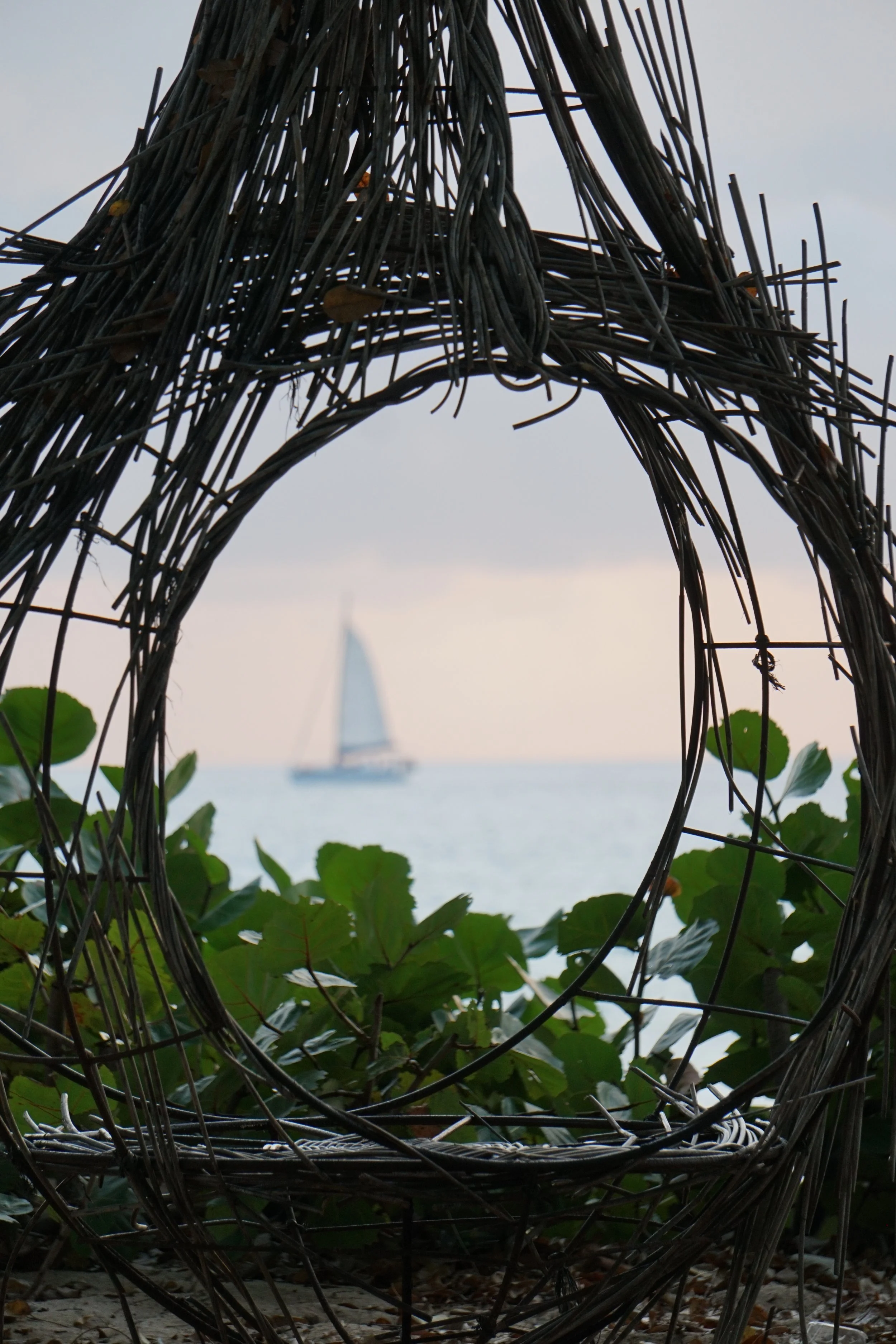 Wicker art sculpture framing view of a sailing boat on the ocean with bushes in the foreground.