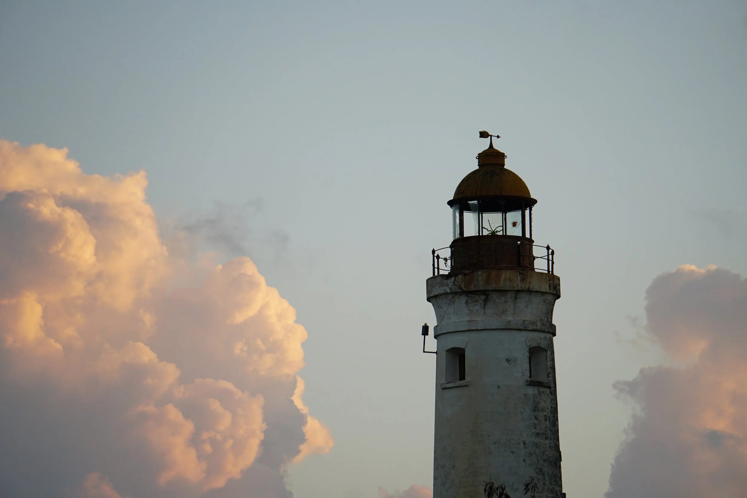 A tall, white lighthouse with a rusted, domed top stands against a sky with large, fluffy clouds illuminated by the setting or rising sun.