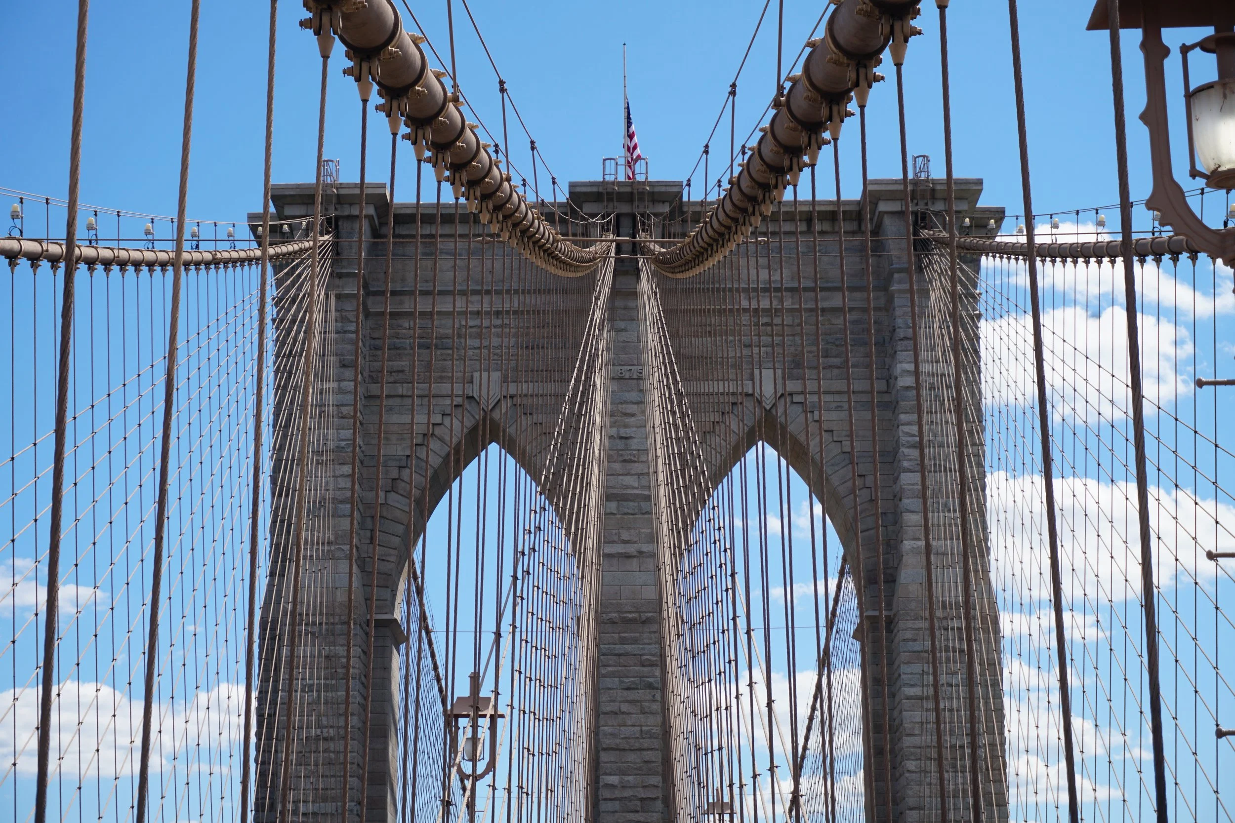 Close-up view of the Brooklyn Bridge's stone towers and suspension cables from the pedestrian walkway with a blue sky and some clouds in the background.