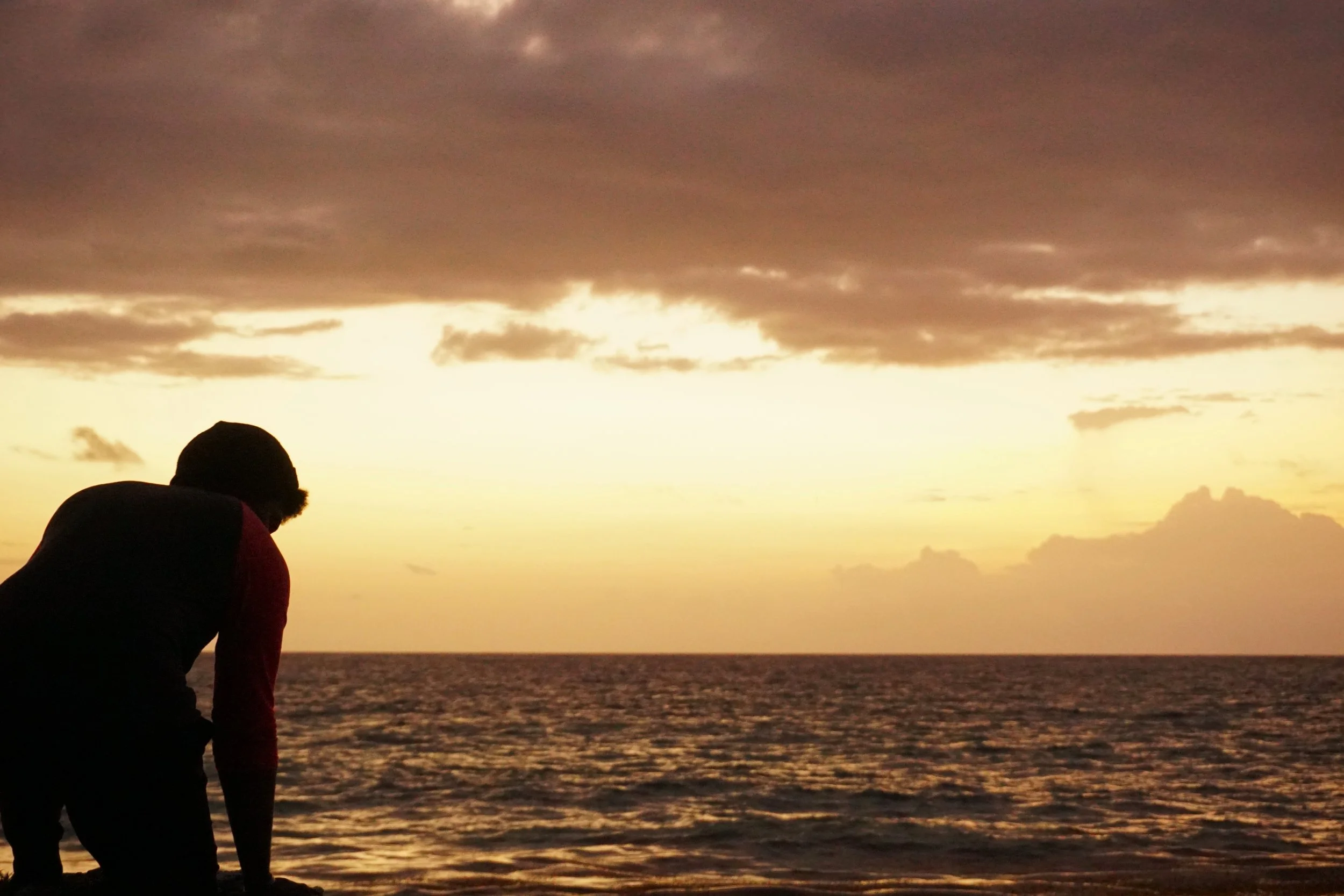 Silhouette of a person near the water during sunset with a cloudy sky.