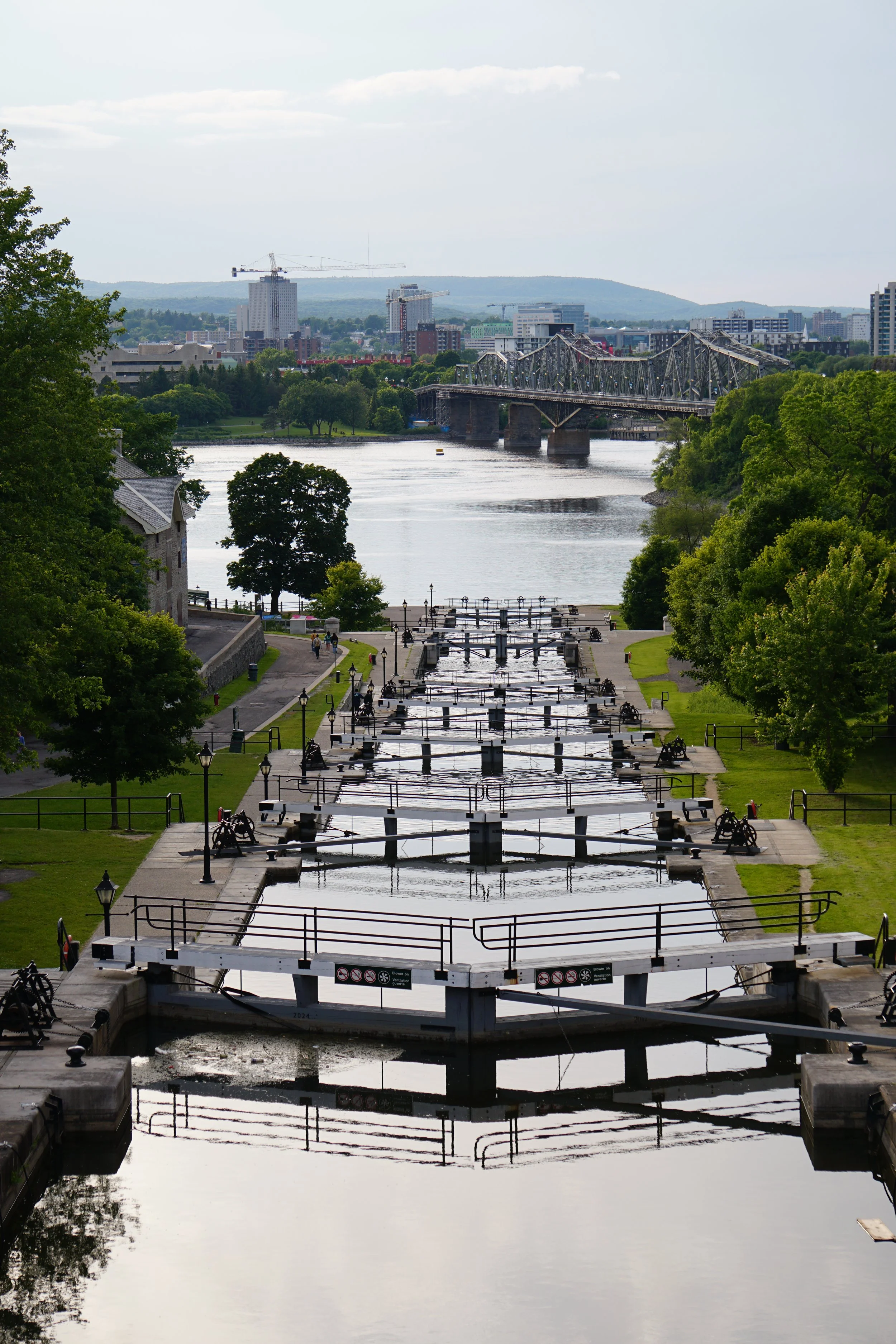 A series of locks on a canal with green trees on both sides and a city skyline with a bridge in the background.
