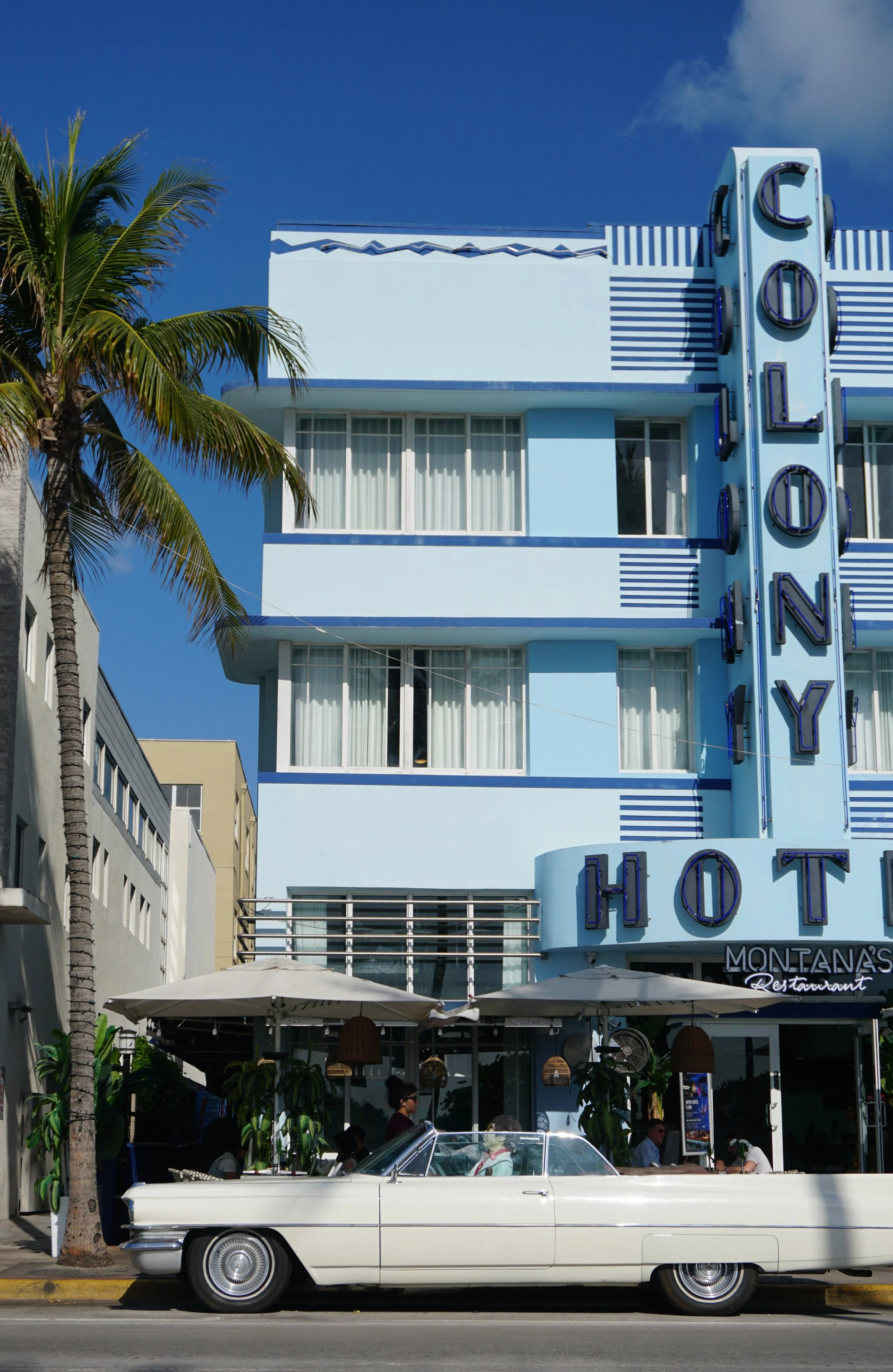 A vintage white convertible car parked in front of a blue hotel building with a large vertical sign that reads 'COLONY' in neon letters, with an outdoor restaurant area underneath shaded by umbrellas and decorated with hanging lamps.