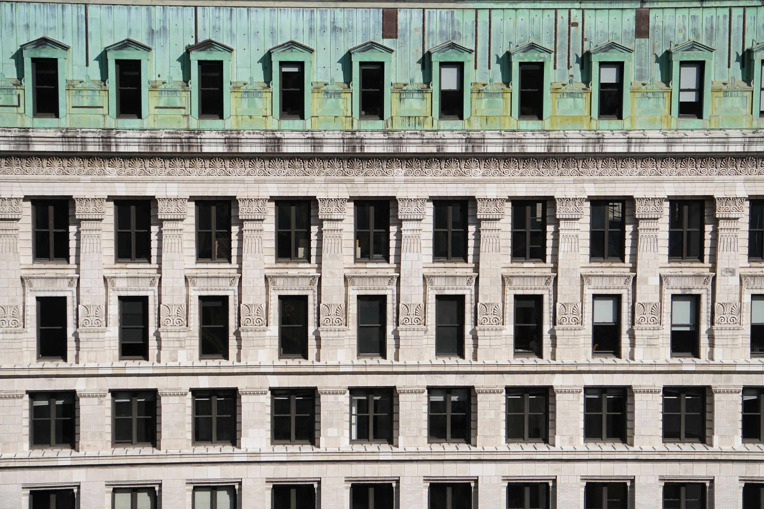 Close-up of a building facade with ornate white stonework and multiple black windows, topped with a green, aged metal roof with small, peaked dormer windows.