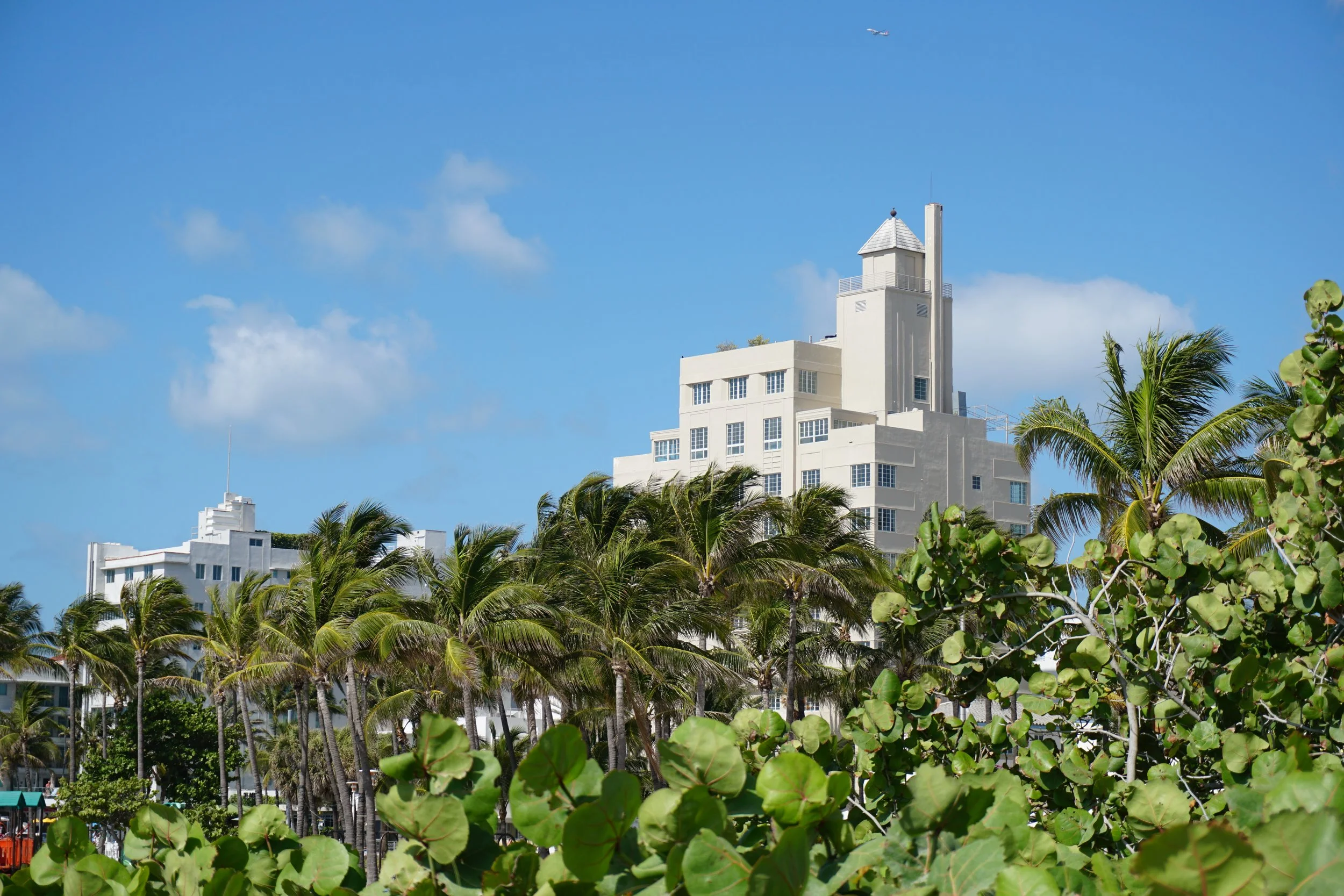 White art deco building behind palm trees and green shrubs with a blue sky and scattered clouds in the background.
