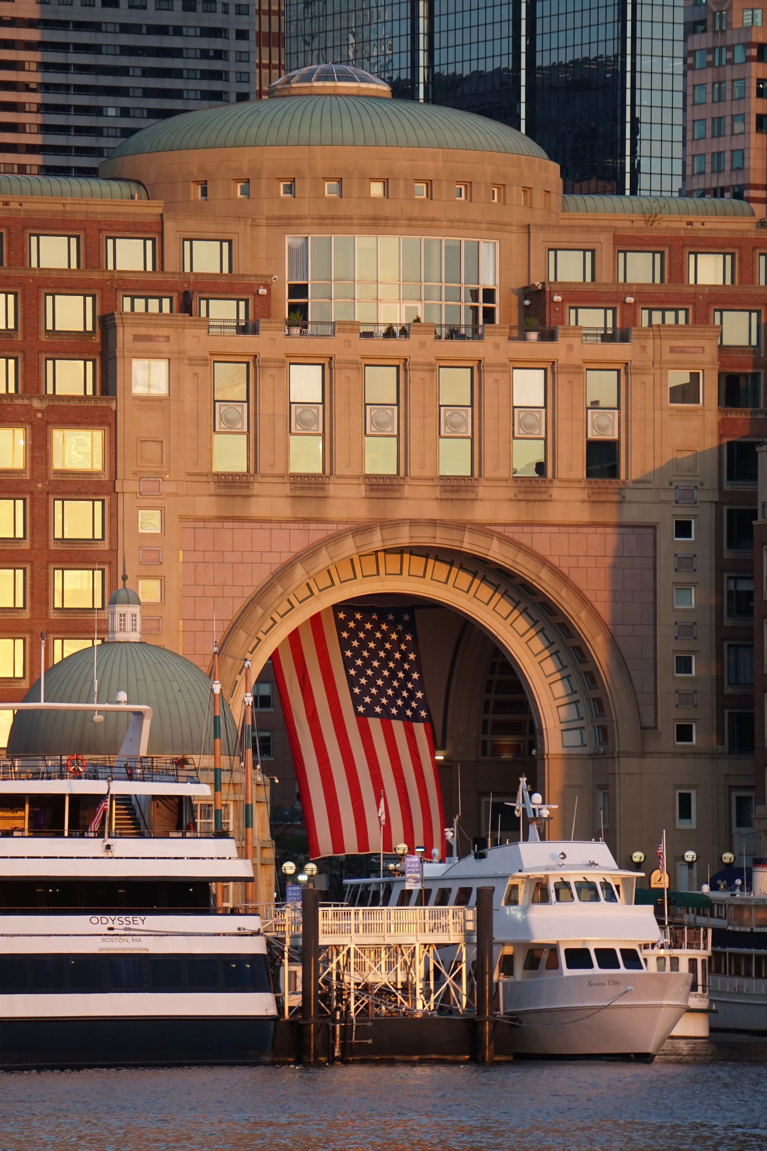 Boats docked at a marina in front of a large building with a grand arch and a suspended American flag, during sunset in Boston, Massachusetts.