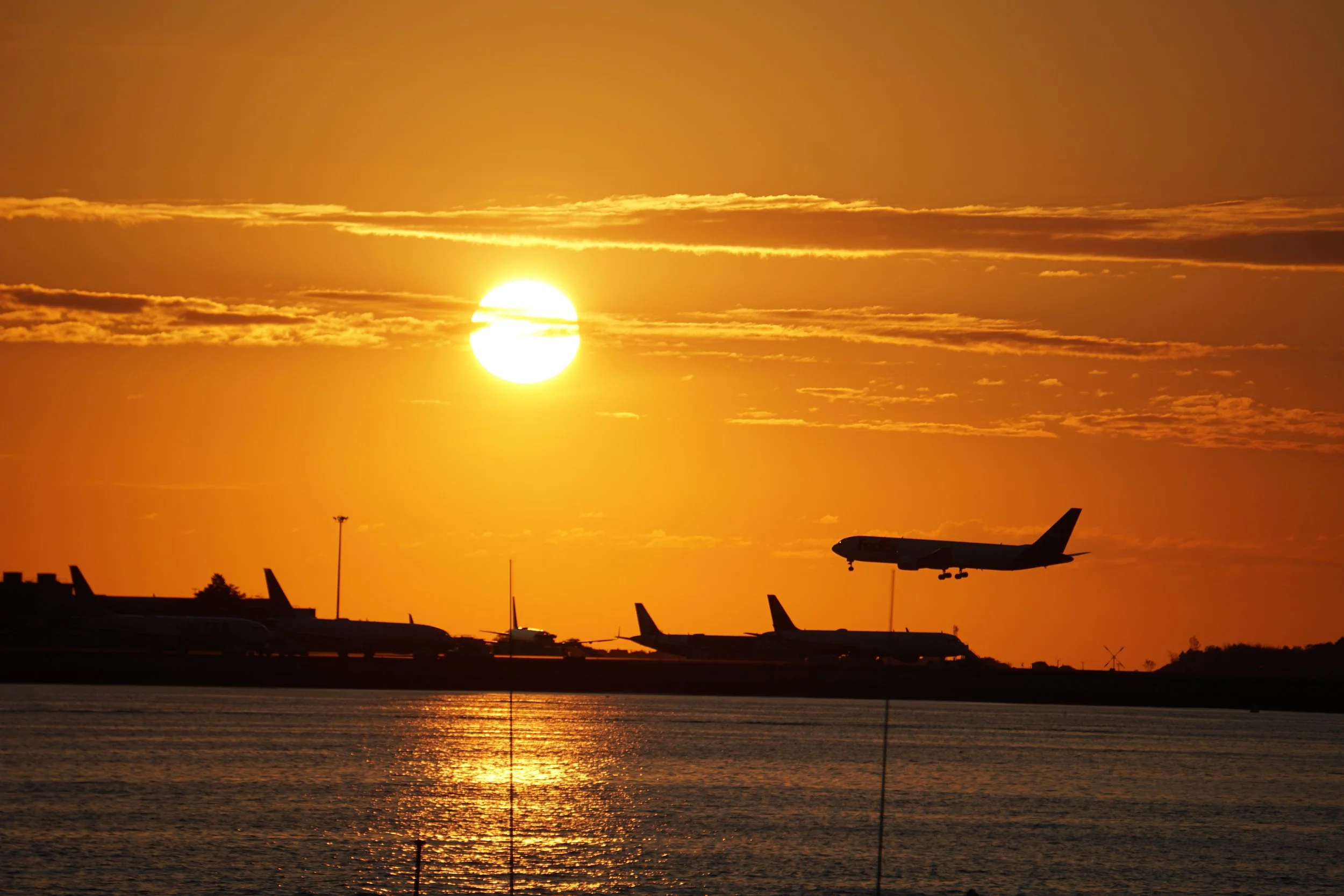 Silhouettes of airplanes at an airport during sunset, with the sun low in the sky and reflecting on the water.