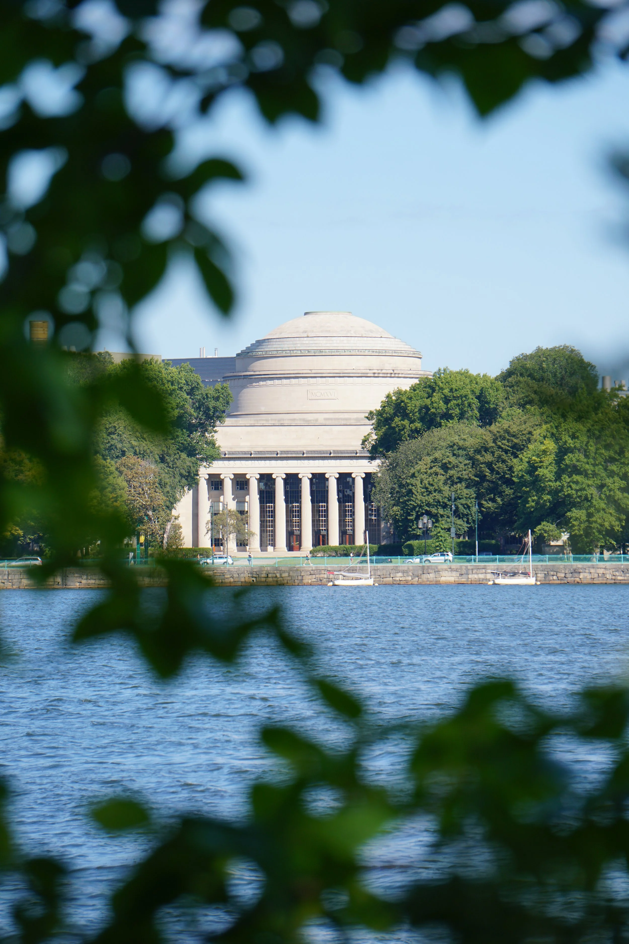 View of a large, domed neoclassical building with columns, seen through leafy branches, across a body of water with two sailboats, trees, and a clear blue sky.
