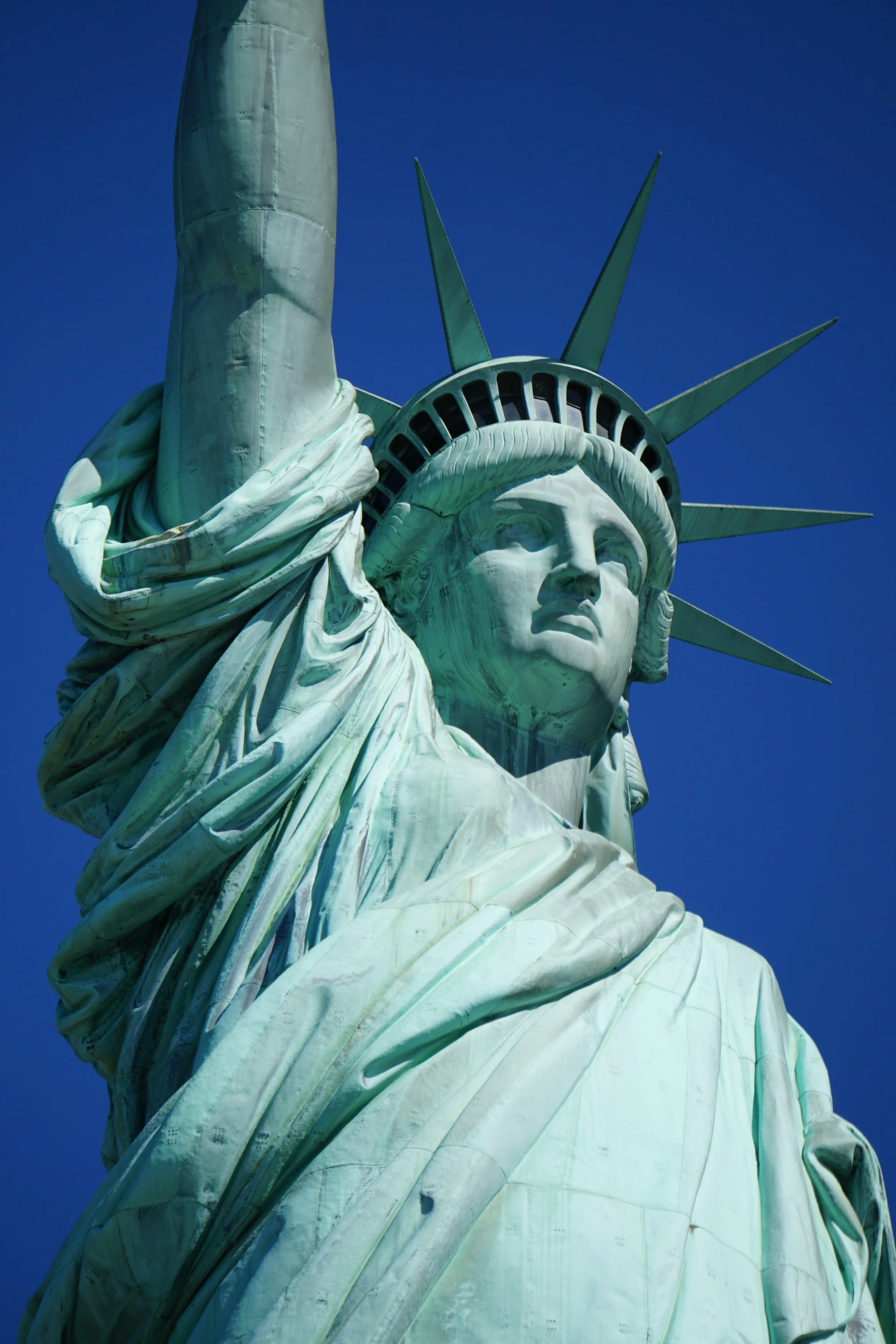 Close-up of the Statue of Liberty's face, crown, and raised arm with a torch against a clear blue sky.