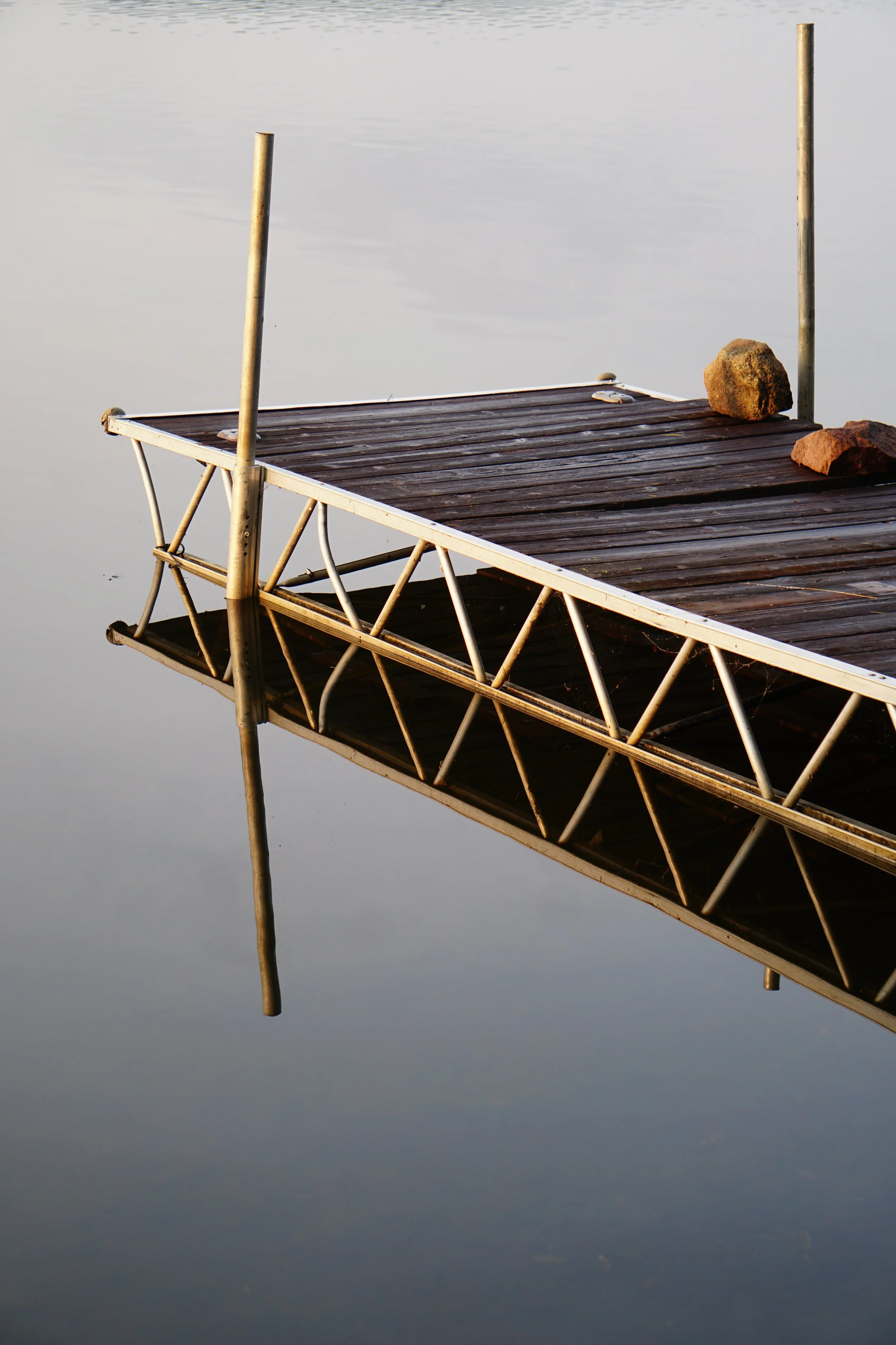 A wooden dock with rocks on it, floating on calm water, with its reflection visible.