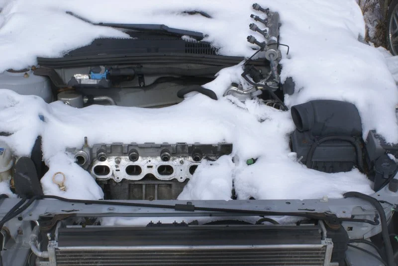 Car engine bay with snow covering parts, missing engine components, and an exposed radiator.