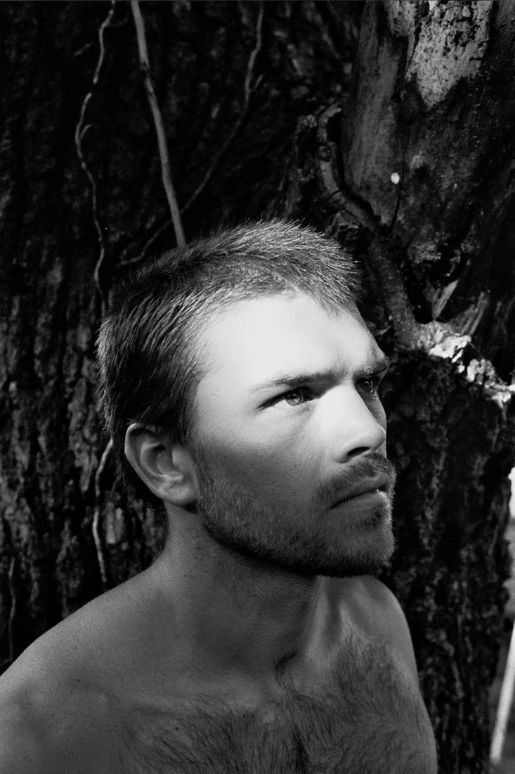 A shirtless man with short hair and a beard looking upward against a background of tree bark.