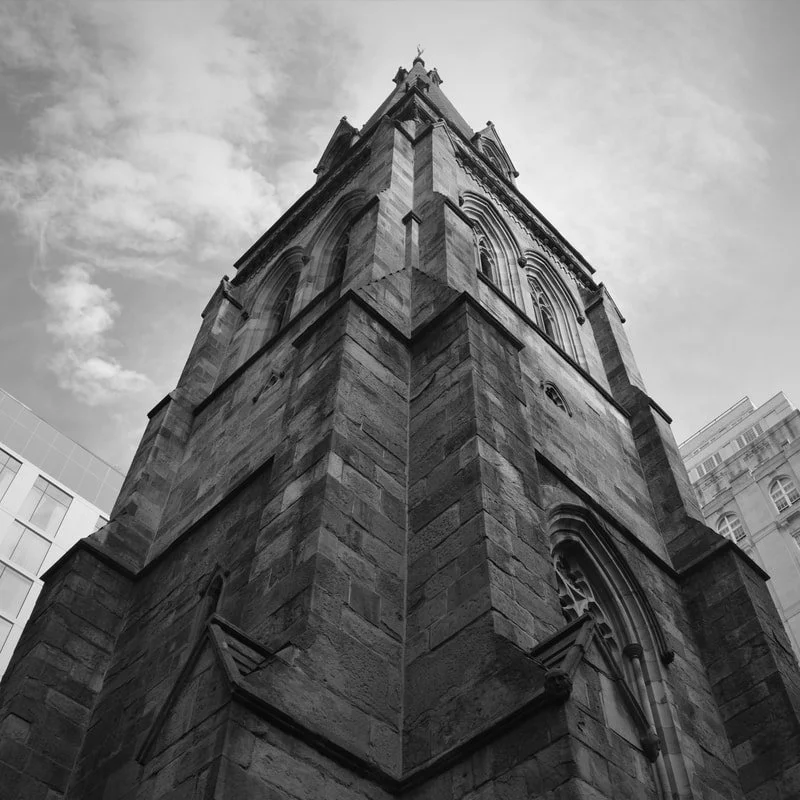 Tall stone church steeple with gothic arches and decorative elements, viewed from the ground up, against a partly cloudy sky.