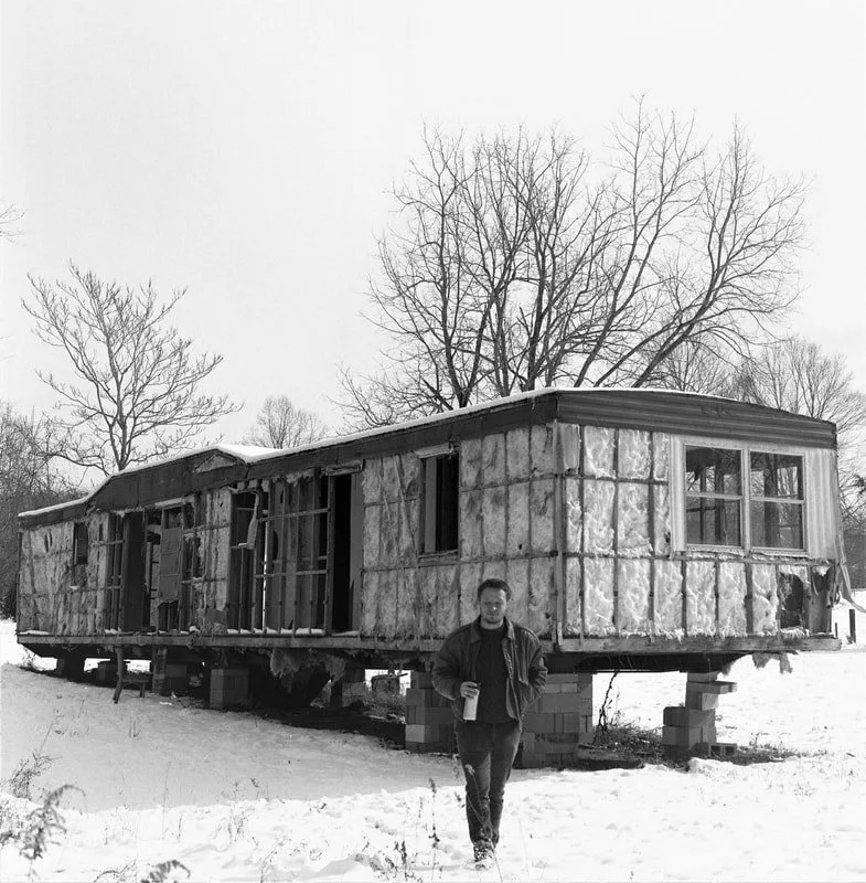 A person standing outdoors in snowy weather holding a drink in front of a mobile home under renovation, with trees and an overcast sky in the background.