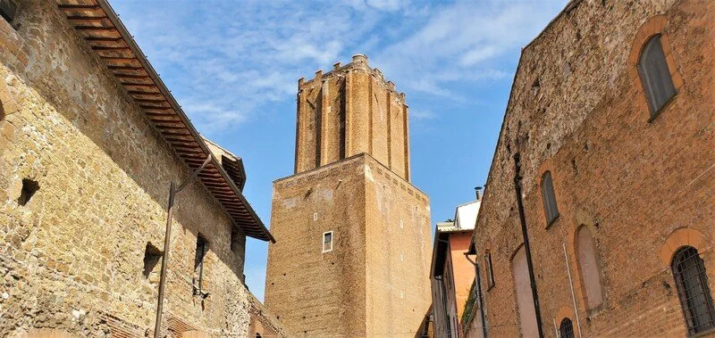 A tall, historic stone tower with a rectangular shape and battlements at the top, surrounded by old brick and stone buildings in a narrow alleyway under a partly cloudy sky.