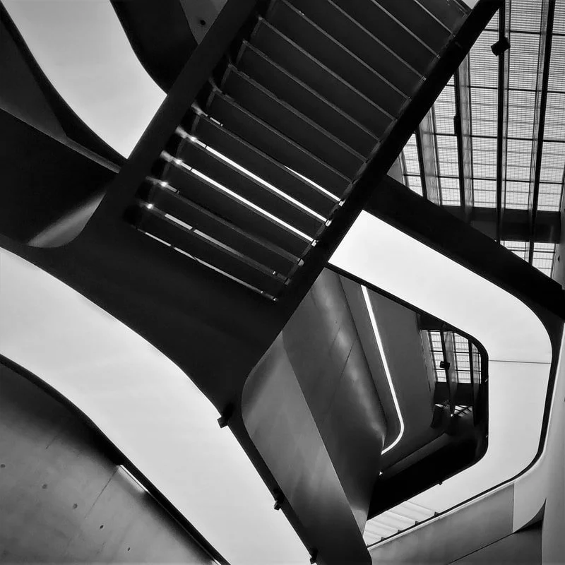 Black and white photo of a modern interior staircase viewed from below, showing sleek, curved lines and glass ceiling.