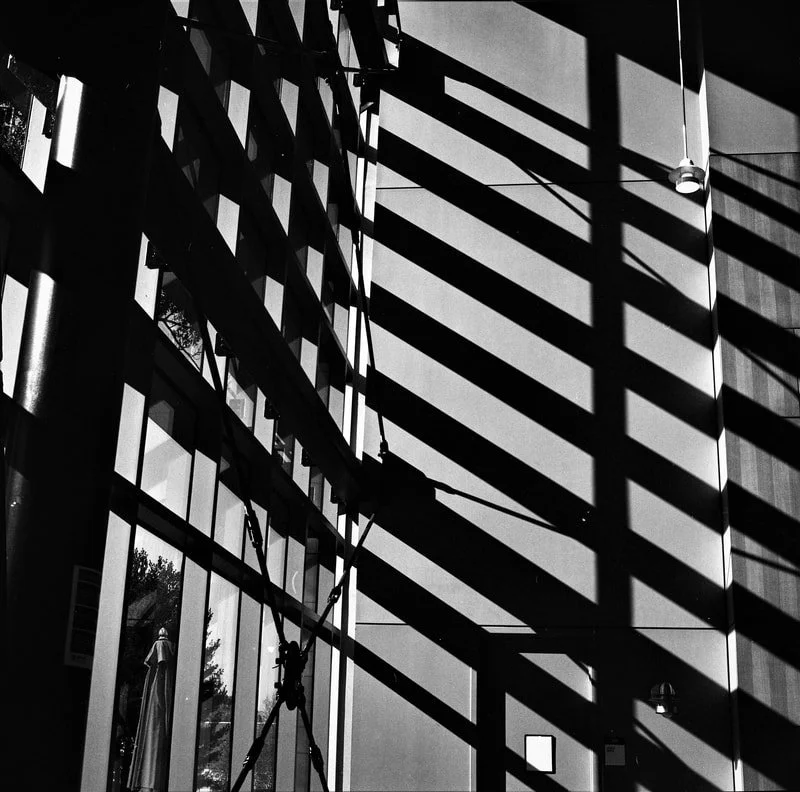 Black and white photo of a building with large glass windows and a metal structure casting shadows of horizontal blinds onto the wall.