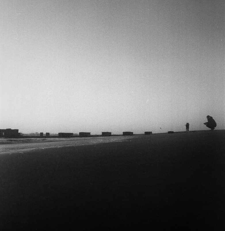 Two people on a beach at sunset or sunrise, one standing and the other squatting, with a row of objects or structures along the shoreline in the distance.
