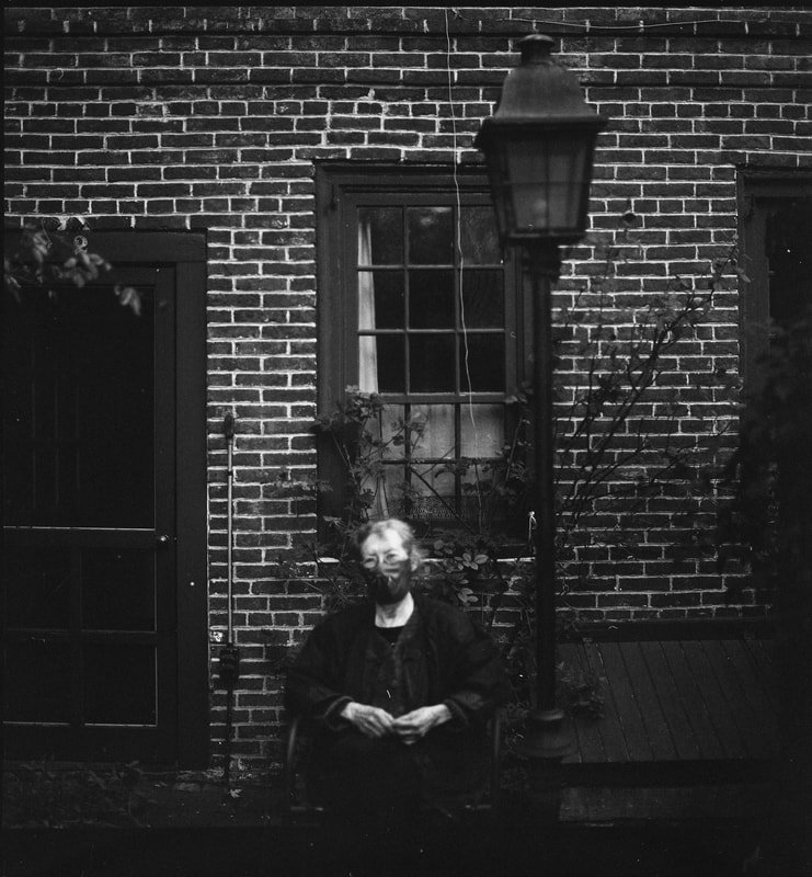 A person sitting outdoors at night in front of a brick wall, smiling, with a street lamp and a window behind them.
