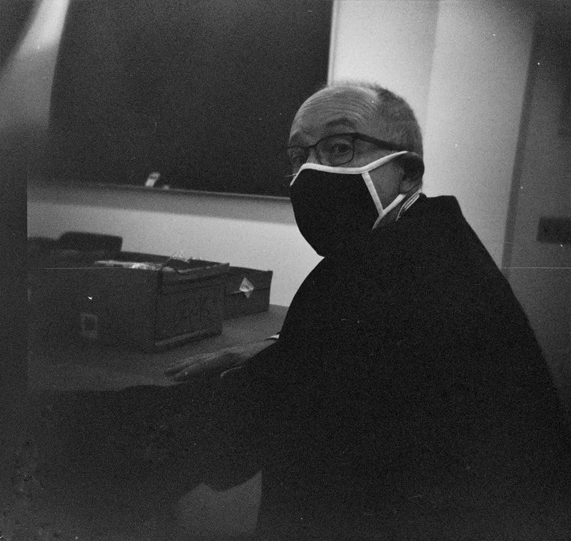 A man wearing glasses and a face mask sitting at a desk in a dimly lit room, with a blackboard and storage box in the background.