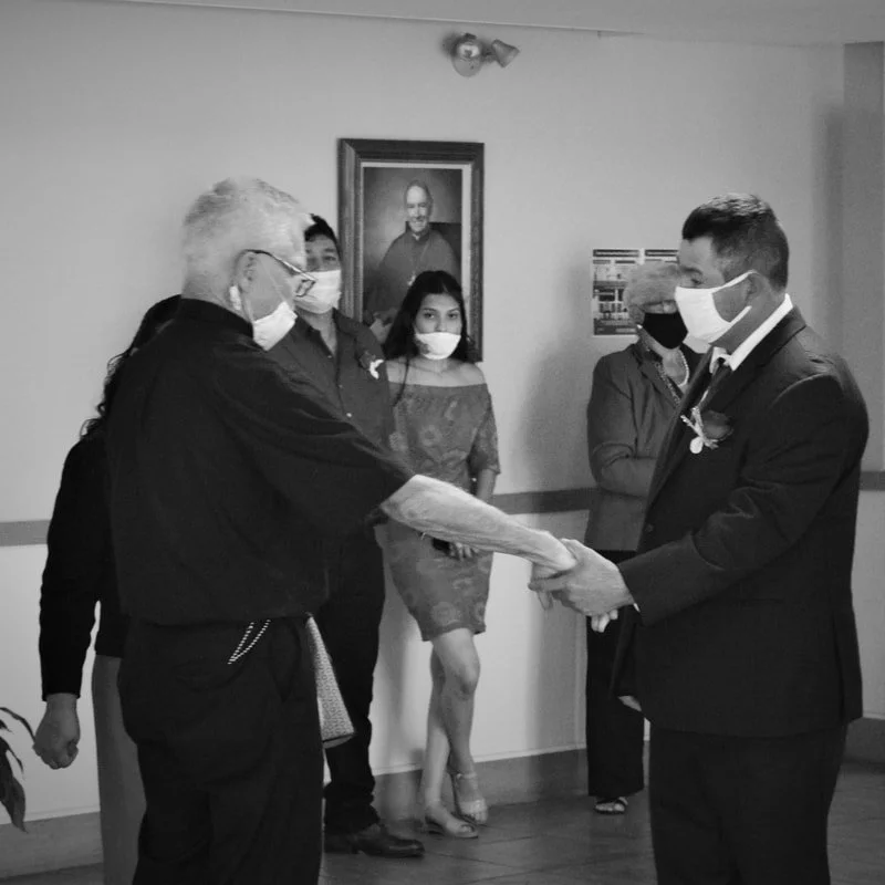 A man in a suit and face mask receiving an award or certificate from an older man with glasses and face mask, with three people in the background, in a room with wall art and a photograph of a man.