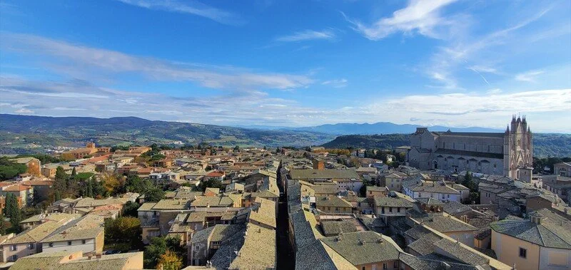 View of a historic Italian city with stone rooftops and a large cathedral in the background under a blue sky.