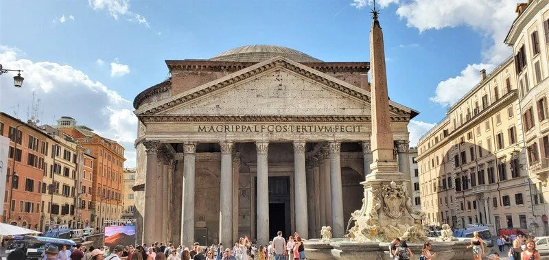 The Pantheon, an ancient Roman temple with a large dome, is surrounded by a crowd of people in a city square. There is an Egyptian obelisk and a fountain in the foreground, with colorful buildings on either side under a blue sky with clouds.