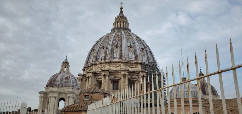 View of a large historic domed church with scaffolding on part of it, surrounded by a rusty iron fence, under a cloudy sky.