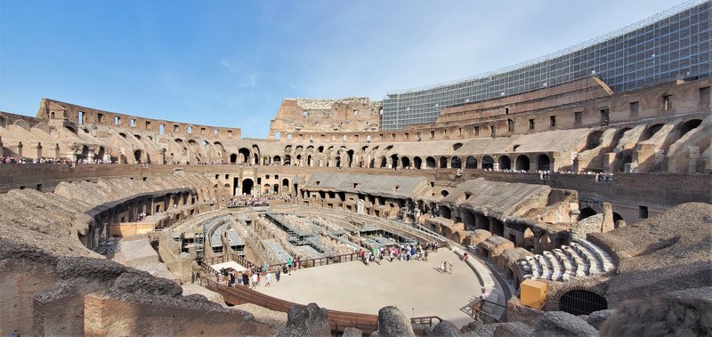 The interior of the ancient Roman Colosseum with tiered seating and a partially excavated arena, under a clear blue sky.