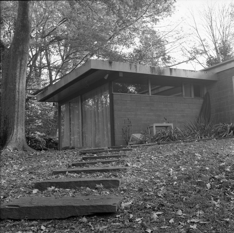 A mid-century modern house with large glass windows, located on a wooded hillside, with steps leading up to the entrance surrounded by fallen leaves.
