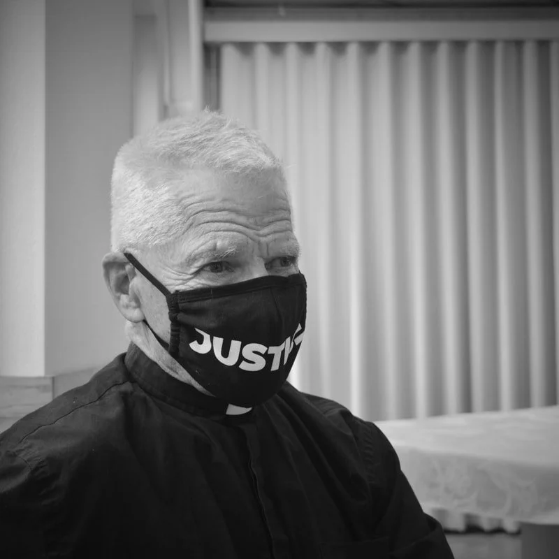 An older man with short, light-colored hair wearing a black face mask with the word 'JUSTICE' on it, sitting indoors near curtains.