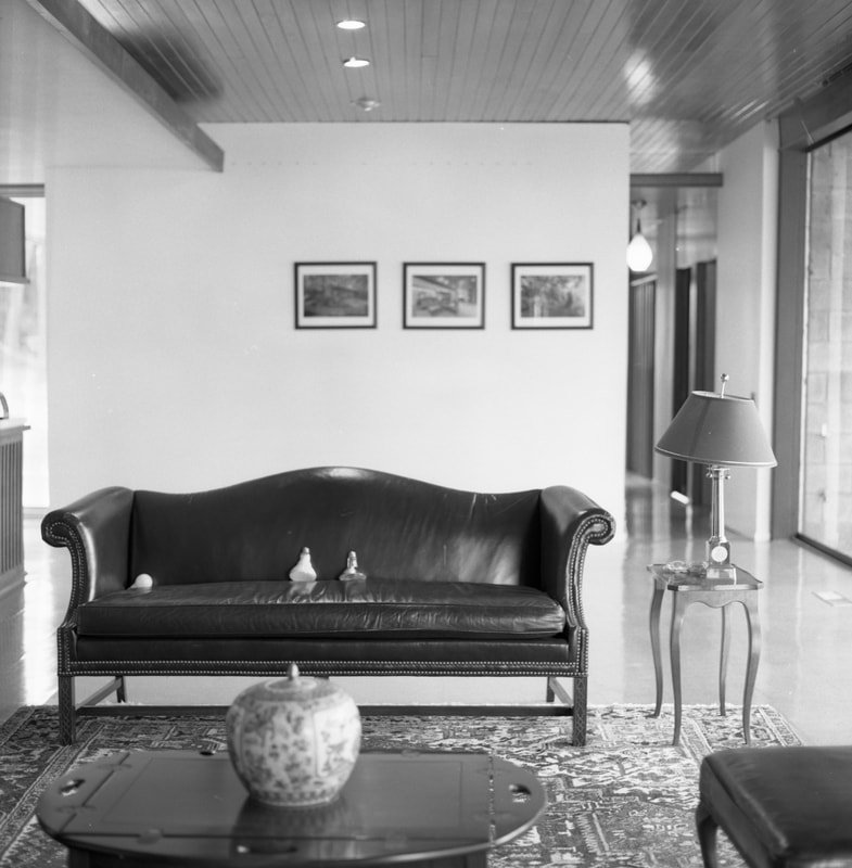 A black leather sofa with decorative nailheads on a patterned rug, a small wooden coffee table with a decorative vase, a side table with a lamp, framed pictures on a white wall, and a glass door or window to the right letting in natural light.