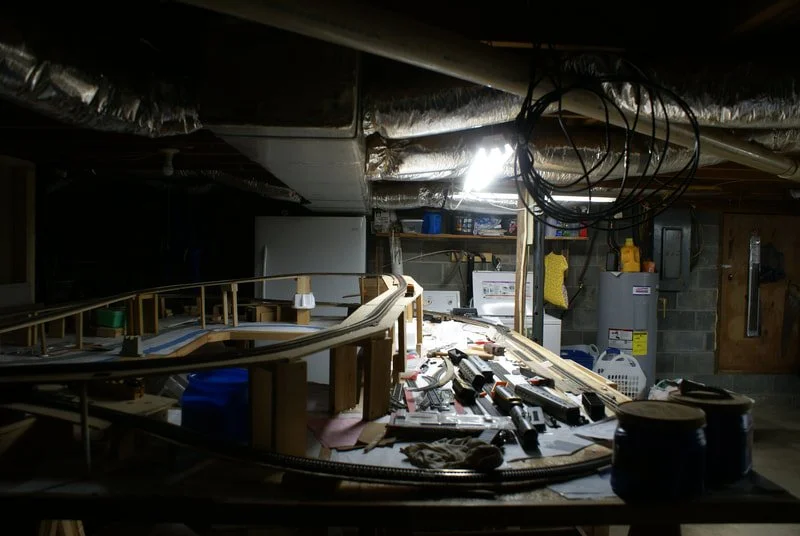 A workbench in an unfinished basement with a model train set, tools, and supplies on it, surrounded by exposed ductwork and clutter.