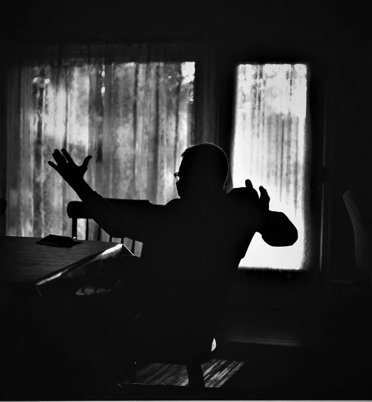 Silhouette of a person sitting at a table with arms raised, in front of a window with curtains, in a dimly lit room.