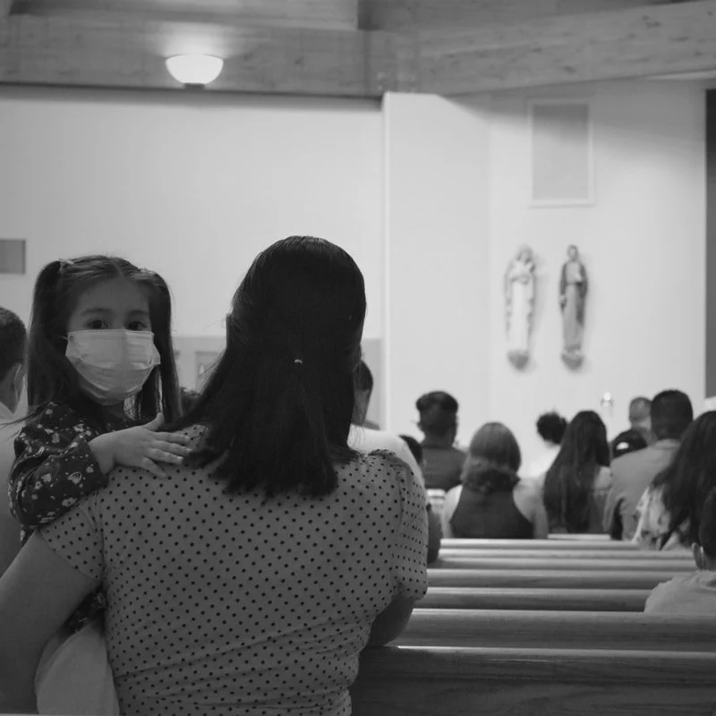 A young girl wearing a face mask sits on a woman's lap during a church service, with an audience seated in front of the altar.