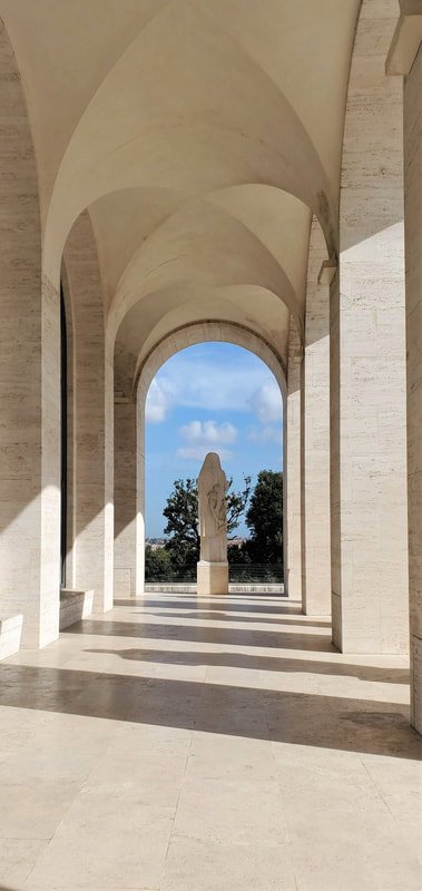 View of a classical architectural structure with arches and columns, featuring a statue in the center and a blue sky with clouds in the background.