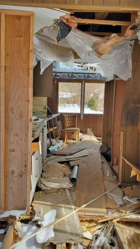 A severely damaged room with a broken ceiling and debris scattered on the floor. A window shows an outdoor snowy landscape. There is a chair and various objects in disarray.