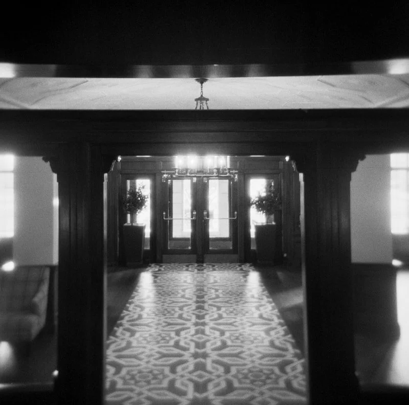 Black and white photo of an elegant lobby with double glass doors, large windows, and potted plants, featuring a patterned carpet and chandelier.