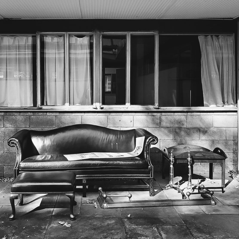 Black leather sofa with matching ottoman and small side table on a porch with concrete floor, in front of a brick wall with large windows covered by curtains.
