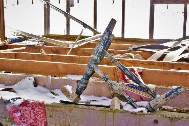 A damaged wooden fence inside a snow-covered windowed room, with fallen and broken wooden beams and a tipped-over wooden chair.