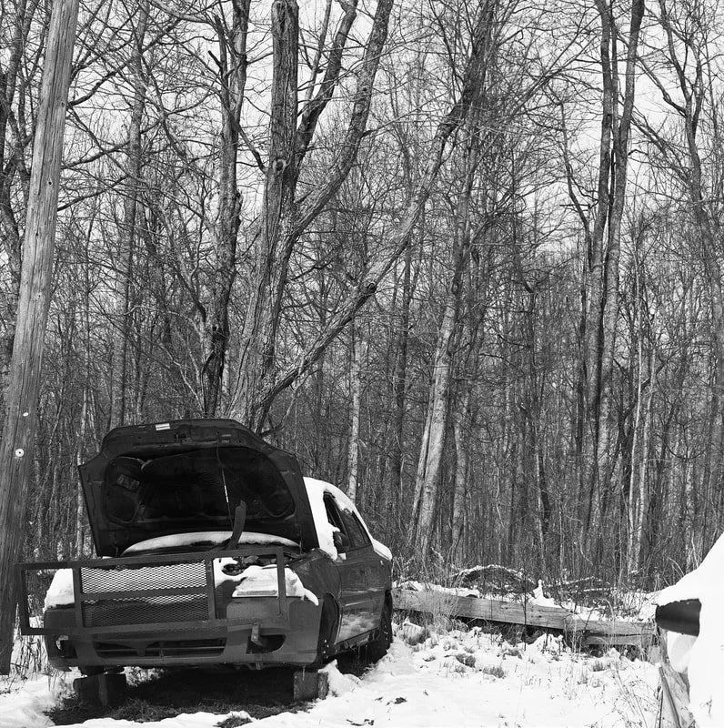A black and white image of a car without front wheels, parked in a snowy forest with leafless trees, with the hood open and a trailer visible on the right side.